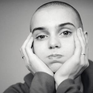 A black-and-white photograph of Sinéad O’Connor in ‘Nothing Compares.' Her head is shaved and she is wearing a long-sleeve shirt. She is resting her head in both of her palms with her fingers clasped over both cheeks. 