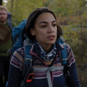 Foreground of image shows Rep. Alexandria Ocasio-Cortez hiking in a green forest. She wears a geometric sweater and a blue backpack; her boyfriend is pictured hiking behind her, slightly out of focus