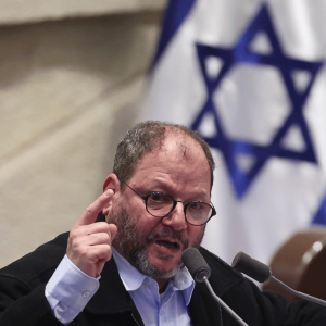 The image shows an older man in a suit and glasses holding up a finger emphatically while speaking in to a microphone. There is an Israeli flag behind him. 