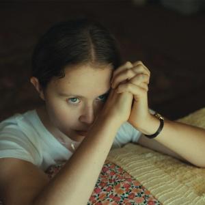 A young white teenage girl named Jem Starling (played by actress Eliza Scanlen) is sitting on the edge of a bed. Here elbows rest on the quilt blanket with her hands folded in prayer as she looks beyond the frame toward an unseen ceiling.