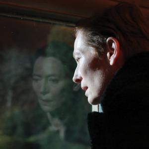 Julie from 'The Eternal Daughter' (Tilda Swinton) is seen from a side profile staring out a window, where you can see her image reflecting in the glass and a view of a forest in the background.