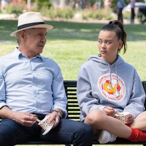 Paul (Harrison Ford) wears a light blue dress shirt and hat and sits on a black park bench next to teenage girl Alice (Lukita Maxwell), who's sitting cross-legged while wearing a gray hoodie.