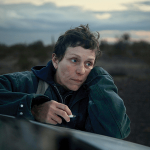 A scene from Nomadland features a woman leaning on the hood of her car.