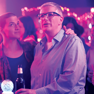 A scene from 'Work In Progress' shows Abby, the main character, at a club in pink light.