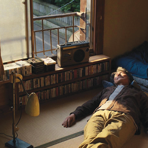 A man is lying down on the floor of his room on tatami mats, with a cassette player and several cassettes neatly organized under an open window.