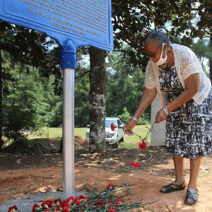 A senior Black woman stoops to lay orange flowers at the base of a plaque