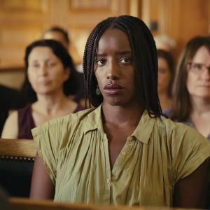 Kayije Kagame plays as Rama in the film ‘Saint Omer.’ She is a Black woman with box braids wearing a creased linen olive-green v-neck dress. She sits in the pews of a court with a crowd of people blurred in the background.