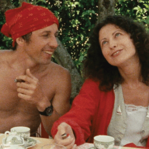 The image shows a man and a woman at a table, laughing about something. The man is shirtless with a red bandana on his head, and the woman has a red shawl thing and dark hair. 