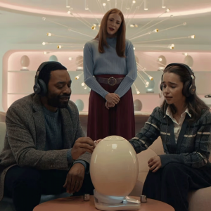 The image shows a couple sitting dow, wearing headphones, looking at a large, electronic egg. There is a woman standing behind them, watching. 