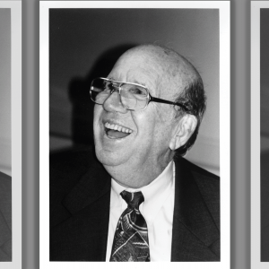 The image shows a black and white photo of a older white man laughing. He is bald and wearing glasses and a suit and tie.