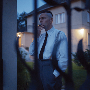 The image shows a Nazi commandant smoking in his yard, and the photo was taken through bars on a fence. The man wears a white button up with a black tie. 