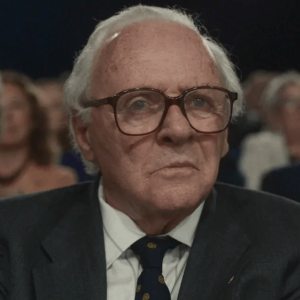 The picture shows an old white man with glasses wearing a suit looking at something, semi-puzzled or surprised. There are people behind him. 