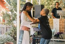 A black woman in a white dress with long dark brown hair smiles while dancing with her elderly mother on a porch, who's wearing a black t-shirt and jeans. A man in a black t-shirt plays music in the background with a laptop and speaker on a table.