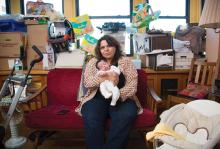 A woman with brown curly hair and a cardigan is sitting on a couch and holding her baby, who has dark hair and wears a white longsleeve footie onesie. Piles of boxes, baby supplies, and furniture surround her on all sides.