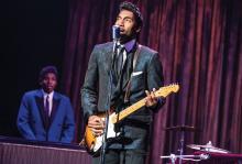 Garrett Turner is dressed in a black suit playing an electric guitar as Ike Turner in the musical 'TINA: The Tina Turner Musical.' A black man in a blue suit is playing the keyboard in the background, where both men are flanked by a purple stage curtain.