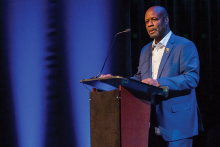  A Black man in a royal blue suit with a white collared shirt stands at a podium; background of the image is dark, with two vertical stripes of blue light extending top-to-bottom to the left of the man.