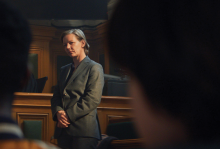The photo shows a woman with gray hair in a gray suit in a courtroom, looking at people off camera. 