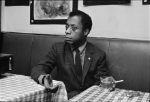 James Baldwin holds a cigarette in one hand and gestures with the other while sitting in a booth