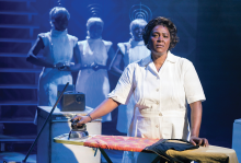 A weary middle-aged Black woman looks up from ironing clothes in a dark room