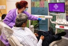 A side-rear view woman doctor with red hair points to a screen with a spectrum of faces from sad to happy, asking her patient in the chair (a man with gray hair) which is most accurate for him. A purple screen with "Church Health" is shown nearby.