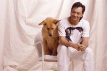 A photo of Rich Mullins in a white t-shirt and white pants. He's sitting on a white chair with a white sheet draped in the backdrop. He's leaning forward on his knees with a grin on his face. A dog with tan-colored fur sits to his right on the same chair.