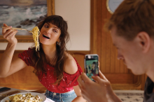 A woman is pictured holding up a fork full of noodles to her smiling mouth while posing for a photo