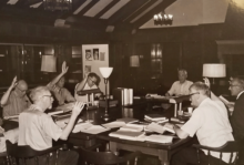 The image is a black and white photo showing a group of old white men sitting around a table with Bibles and other documents, some have their hands raised. 