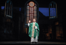 The image is a picture of a priest on a stage with a green robe, with a stained glass window in the back