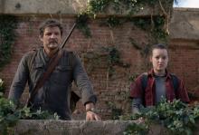 Joel (actor Pedro Pascal) and Ellie (actress Bella Ramsey) from 'The Last of Us' HBO series are standing side by side on the roof of a neglected building with their arms leaning on a brick wall covered in foliage.