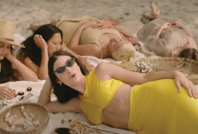 Lorde, wearing yellow, sings to the camera as she lays on a blanket on the sand
