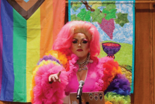 A person wearing a tall pink wig and a pink dress with rainbow fluffy sleeves is standing at the pulpit of a church, preaching. There is a pride flag in the background. 