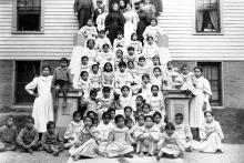 An old black-and-white photo of students and teachers sitting and standing on the steps of the Thomas Indian School building in the 1890s. 