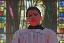 A young boy wearing a white surplice stands before a wall of stained glass