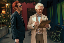 The photo shows two men, one who is an angel and dressed in lighter colors, and another who is a demon dressed in black. The angel is looking at a clipboard and the demon is just standing there. 