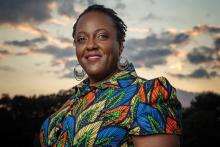 A photo of Heather McTeer Toney: a black woman with short hair, golden circular earrings, and a shirt with a pattern of leaves in vibrant blues, oranges, and yellows. She is looking at the viewer and smiling with a forest and evening sky behind her.