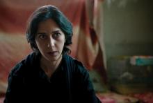 A photo of actress Zar Amir Ebrahimi as fictional journalist Arezoo Ramimi in the film 'Holy Spider.' She is cast against a red flag in the background and staring just off camera at something.