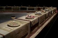 A picture of Judy Chicago's art exhibit called "The Dinner Party." There are unique plates, glasses, silverware, and tapestries depicted for esteemed women across time (mythological and historical) around a triangular banquet table. 