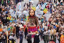 An 11-foot puppet designed to look like a Syrian child is surrounded by a crowd with signs advocating for relief for refugees.