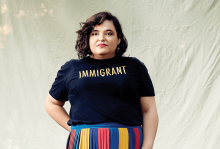 Aline Mello stares seriously at the camera wearing a multicolored skirt and a t-shirt that says "Immigrant"