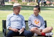Paul (Harrison Ford) wears a light blue dress shirt and hat and sits on a black park bench next to teenage girl Alice (Lukita Maxwell), who's sitting cross-legged while wearing a gray hoodie.