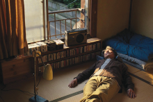 A man is lying down on the floor of his room on tatami mats, with a cassette player and several cassettes neatly organized under an open window.