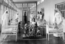 Black and white image of nuns in a "mother and baby home" line a room filled with children in several cribs