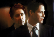 The image shows a scene from "The Devil's Advocate," where one white man is looking over the shoulder of another white man in a suit, who is looking out a window. 