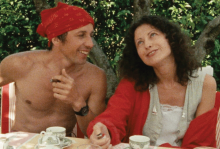 The image shows a man and a woman at a table, laughing about something. The man is shirtless with a red bandana on his head, and the woman has a red shawl thing and dark hair. 