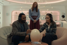 The image shows a couple sitting dow, wearing headphones, looking at a large, electronic egg. There is a woman standing behind them, watching. 