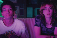 The image shows two teenagers bathed in pink/purple light staring at something behind the camera.