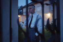 The image shows a Nazi commandant smoking in his yard, and the photo was taken through bars on a fence. The man wears a white button up with a black tie. 