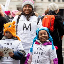 A woman and two children stand in a crowd, wearing white t-shirts that say "You Raise I Rise" 