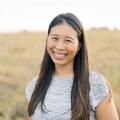 The image shows a smiling young Asian woman in a field. She is wearing a gray shirt. 