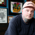 The image shows a white man wearing a hat that says "Grateful" looking at the camera. He has neck tattoos. 